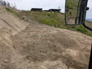 Final grading of a uniform distribution sand mound septic system in a wildfire recovery zone, featuring accessible green inspection lids and a view of modern hillside homes in the Okanagan