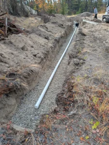Installation of PVC piping on a gravel bed within a long standard gravity septic trench, featuring manual grading work in a wooded Okanagan area.