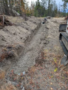 Backfilling a standard gravity septic trench with drain rock to protect the perforated pipe, showing manual spreading of gravel in a residential installation in the Okanagan
